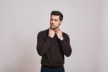 He cares about his style. Young dark-haired man wearing brown jumper standing over grey background