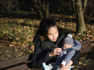 Naklejka na ściany i meble Happy Asian young mother walking with baby outdoors in the park during Autumn, good cold weather, happy child, mixed race family, half Asian Half European. Autumn scene of mother kissing young baby
