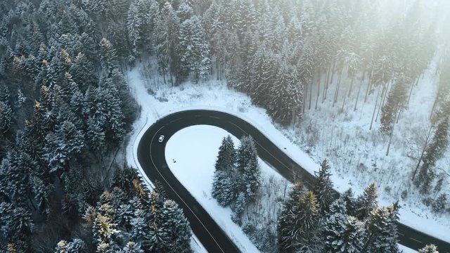 Overhead aerial top view over car travelling on hairpin bend turn road in mountain winter snow covered forest. White pine tree woods. Snowy street path establisher. 4k drone flight sidewards