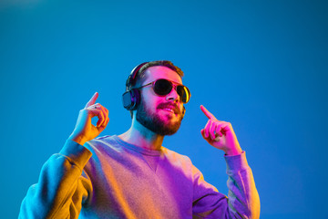 Enjoying his favorite music. Happy young stylish man in hat and sunglasses with headphones listening and smiling while standing against blue neon background