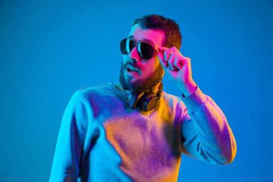 Enjoying his favorite music. Happy young stylish man in hat and sunglasses with headphones listening and smiling while standing against blue neon background