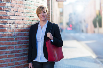 Front view of a confidence, elegant and smiling business woman leaning on a building wall while looking camera outdoors in sunny day