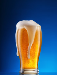 Glass of light beer with drip foam on blue background
