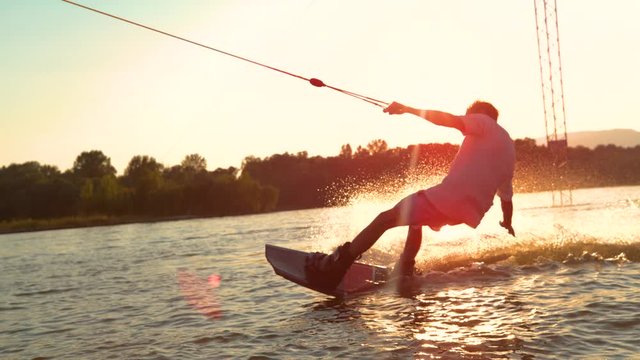 SLOW MOTION, CLOSE UP, SUN FLARE: Unrecognizable athletic man wakesurfing on the lake at golden sunset. Young surfer dude having fun wakeboarding in the cable park on a beautiful summer morning.