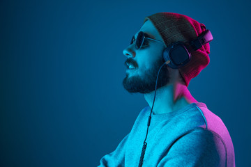 Enjoying his favorite music. Happy young stylish man in hat and sunglasses with headphones listening and smiling while standing against blue neon background