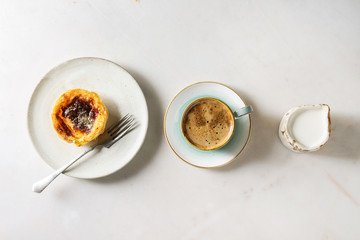 Traditional Portuguese egg tart dessert Pasteis Pastel de nata on ceramic plate with fork, cup of black coffee and jug of cream in row over white marble background. Flat lay, space