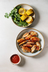 BBQ spicy chicken wings served in ceramic plate with red sauce, grilled sweet corn and parsley bundle in bowls over white marble background. Flat lay, space.