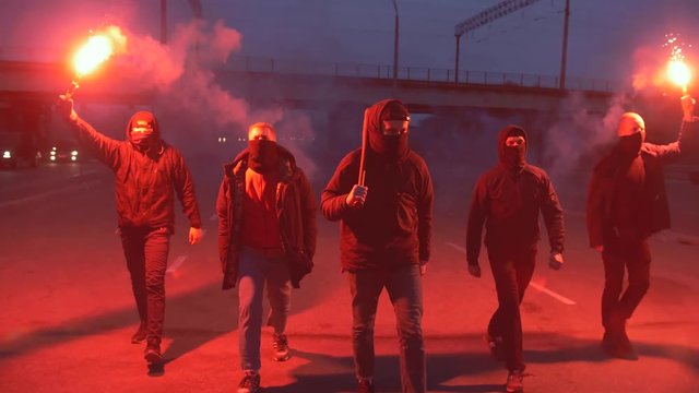 Group of young men in balaclavas with red burning signal flare walking on the road under the bridge, slow motion