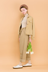 trendy woman in beret holding bouquet isolated on beige