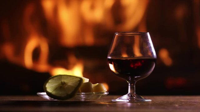 Still life of a glass of brandy and plate with cheese and lemon on the background of a burning fireplace