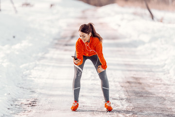 Happy Caucasian sporty woman using smart phone while resting and holding hand on the knee. Winter fitness concept.
