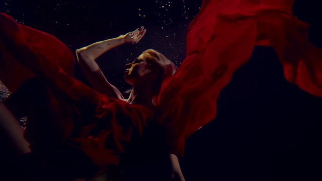 Lady is diving underwater in darkness, wrapped red silk fabric