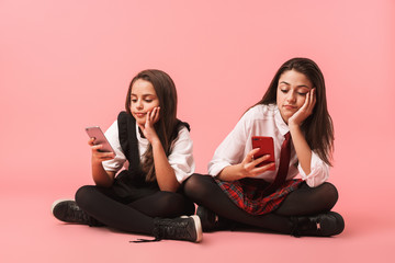 Portrait of beautiful girls in school uniform using mobile phones, while sitting on floor isolated...