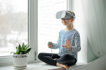 A child with a virtual reality headset is sitting at the table indoors at home. A girl exploring the world of virtual reality through video and games.