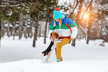 Fototapeta premium Portrait of cheerful young woman in warm stylish clothes and bagpack posing with her beagle dog in winter park. Friendship, pet and human.