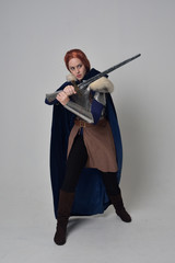 full length portrait of a  red haired girl wearing medieval warrior costume and steel armour and a fur cloak, standing pose on grey studio background.