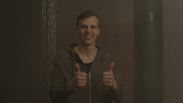 Portrait of cheerful man in hoodie showing thumbs up with both hands in foggy gym. Positive casual guy gesturing tumbs up sign, expressing happiness and joy indoors.
