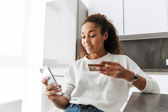 Image of young african american girl using mobile phone and credit card, while sitting at table in bright living room