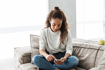 Obraz premium Photo of adorable african american woman using cell phone, while sitting on couch in bright apartment