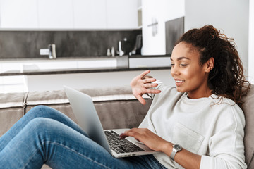 Photo of brunette african american woman using laptop, while lying on sofa in bright living room