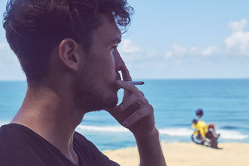 Young handsome man smoking cigar while standing near motorcycle on the tropical beach.