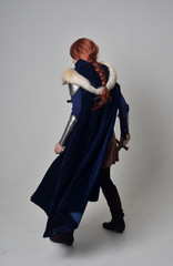 full length portrait of a  red haired girl wearing medieval warrior costume and steel armour and a fur cloak, standing pose on grey studio background.