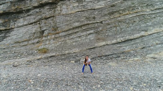 Woman is doing squats the bodyflex on the rock and sea background. Aerial beautiful footage view.