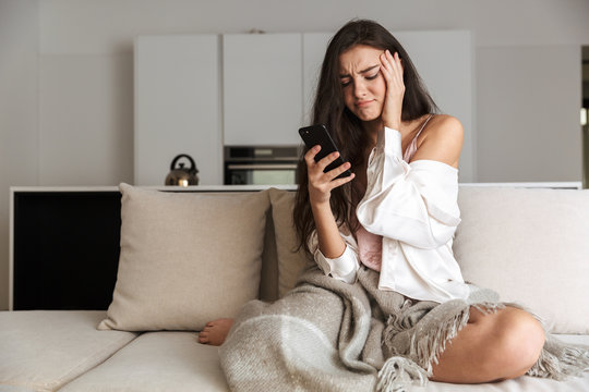 Photo Of Displeased Woman 20s Wrapped In Blanket, Sitting On Sofa At Home And Using Smartphone