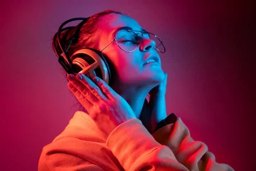 Fototapete Fashion pretty woman with headphones listening to music over red neon background at studio. © master1305