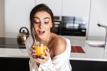 Photo of european woman 20s wearing silk leisure clothing, drinking juice in kitchen