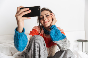 Photo of pretty woman 20s wearing earphones, taking selfie on cell phone in bedroom