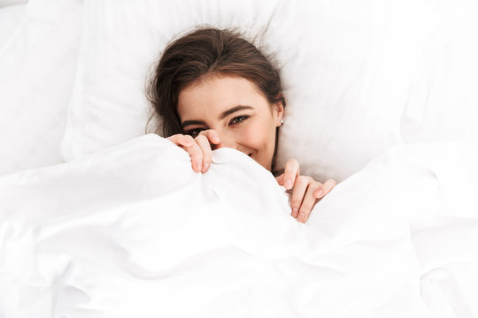 Photo from above of cute woman 20s smiling, while lying in bed on white pillow after sleep or nap