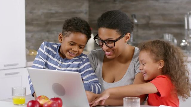 Lockdown of African woman sitting at PC in kitchen with her son and daughter, typing, laughing and then hugging them