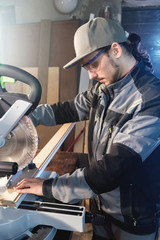 Young brunette man wearing a cap in a gray jacket by profession a carpenter cuts wooden boards with a circular saw on a workbench table in a workshop. Professional equipment in home workshops