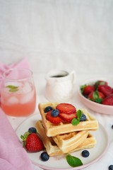 Stack of Homemade fluffy waffles with berries syrup and pink lemonade on warm white background