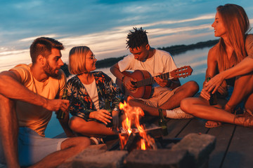 Group of young friends enjoying at the lake at night. They sitting around the fire singing and having fun.