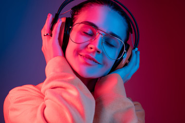 Fashion pretty woman with headphones listening to music over red neon background at studio.