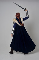 full length portrait of a  red haired girl wearing medieval warrior costume and steel armour and a fur cloak, standing pose on grey studio background.