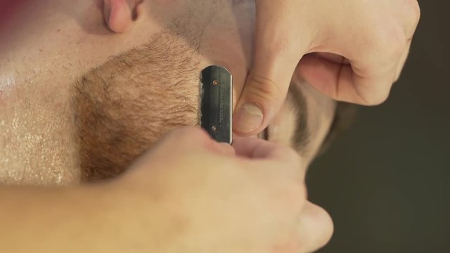 The barber carefully shaves the client's beard using a special shaving razor. It is a close up video of vertical orientation. The background is blurred.