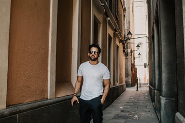Obraz premium Content attractive hispanic man in white tshirt walking old town streets holding mobile phone hand.Bearded hipster male enjoying city travel on gotic district streets