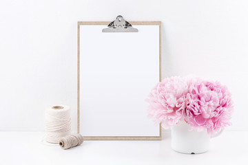clipboard mock up with blank sheet of paper (DIN A4, portrait format), two yarn spools and a bunch of pink peonies on a white table - copyspace for design or text - Powered by Adobe