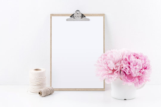 clipboard mock up with blank sheet of paper (DIN A4, portrait format), two yarn spools and a bunch of pink peonies on a white table - copyspace for design or text
