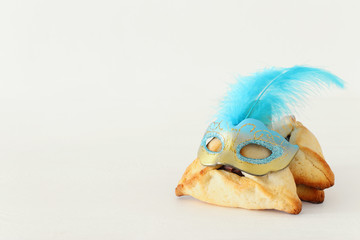 Purim celebration concept (jewish carnival holiday). Traditional hamantaschen cookies with cute mask over white wooden table