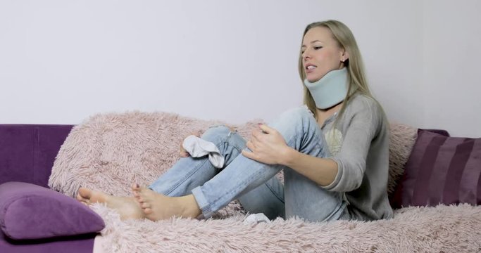 Blonde woman wearing a neck brace sitting on her couch at home falls over trying to put on her socks, gets one on and pauses looking at the other sock in frustration