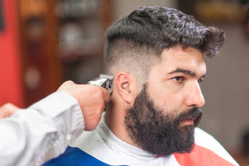 Fototapeta premium Handsome bearded man, getting haircut by barber, with electric trimmer at barbershop .