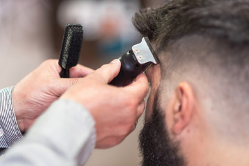 Handsome bearded man, getting haircut by barber, with electric trimmer at barbershop .
