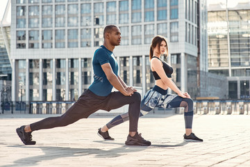 Fototapeta premium Stretching after great workout. Side view of sporty and healthy couple stretching legs during morning workout against modern buildings.