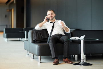 Happy businessman in formalwear having drink and talking by smartphone while relaxing on black...