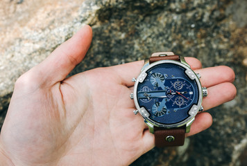 Guy is holding fashionable elegant men's watch in hands. Stylish accessories in the forest on stone. Copy space place.