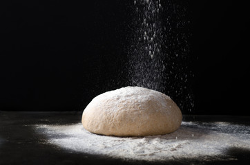 Fototapeta na wymiar Flour pours on fresh homemade dough. Dough with spilled flour on a black background. Yeast dough for bread, rolls, pizza or pie. Copy space.